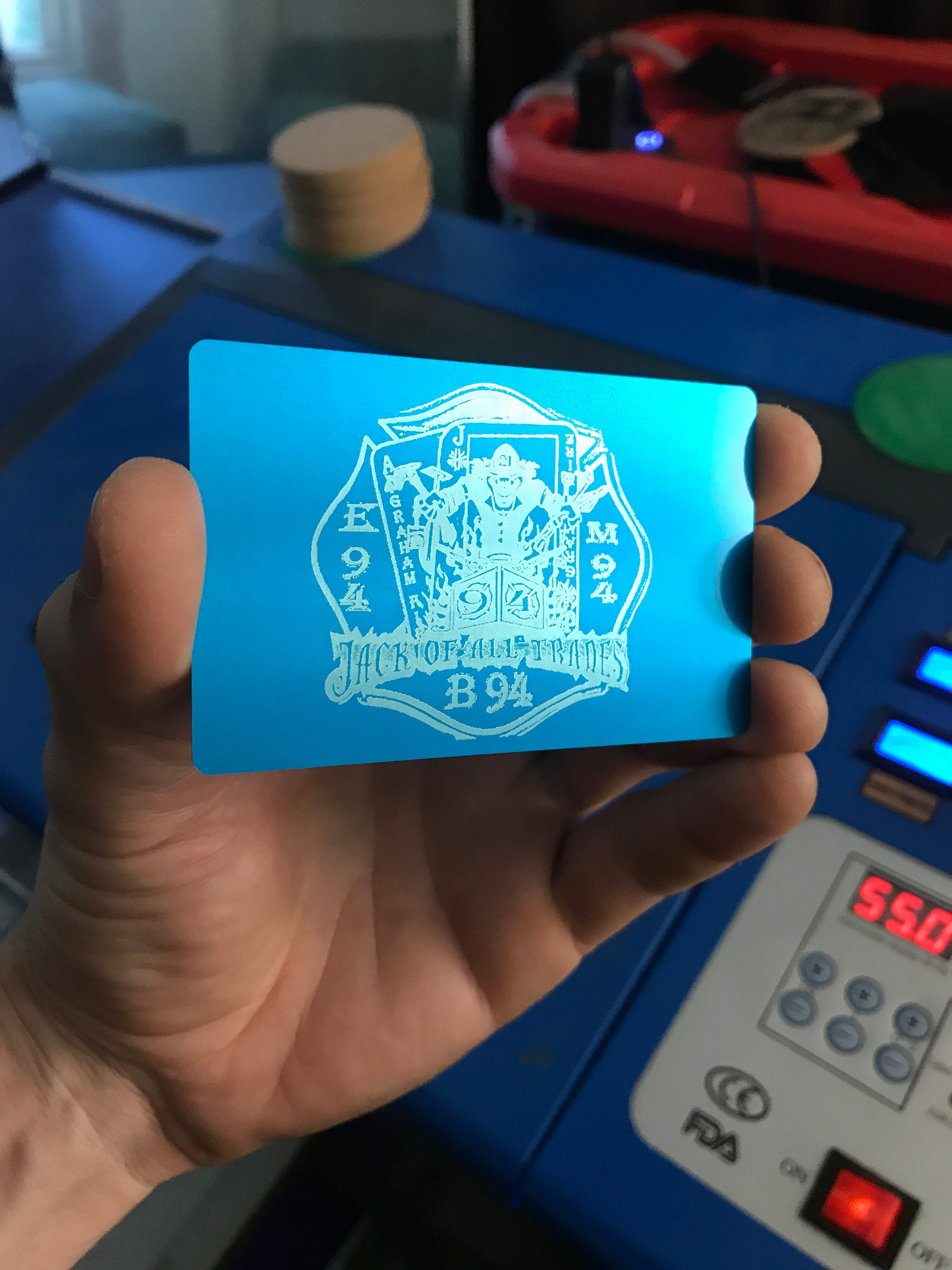 Hand holding a blue/silver laser engraved business card with a customized fire department logo and text in front of a cnc machine with digital display.