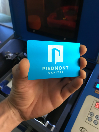 Hand holding a blue and silver metal business card with custom business branding engraved in front of a cnc machine.