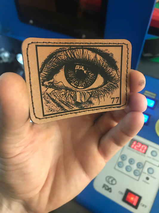Hand holding a leather heat transfer patch with an eye illustration and number 77, against a blurred background.
