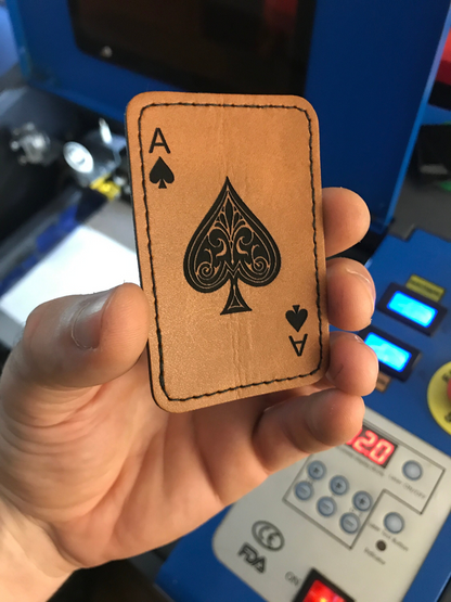 Hand holding a leather patch with an ace of spades design in front of a machine.