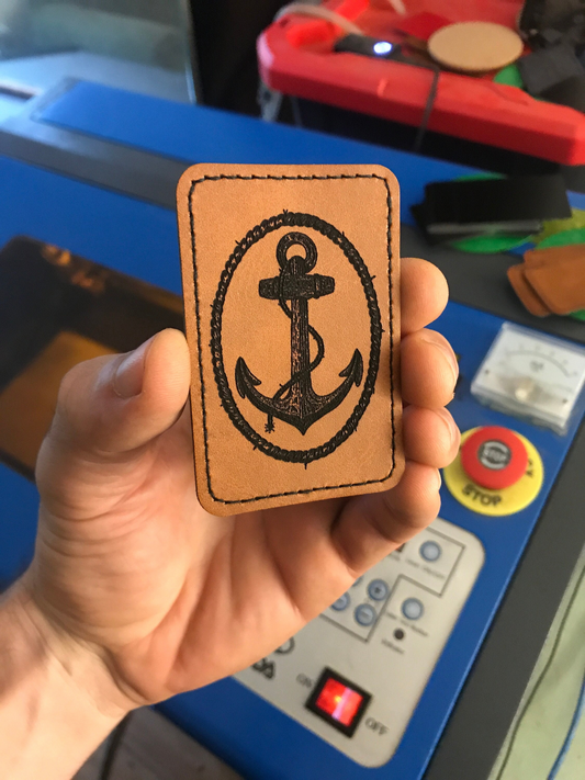 Hand holding a brown leather patch with a rope knot anchor design in front of a machine.
