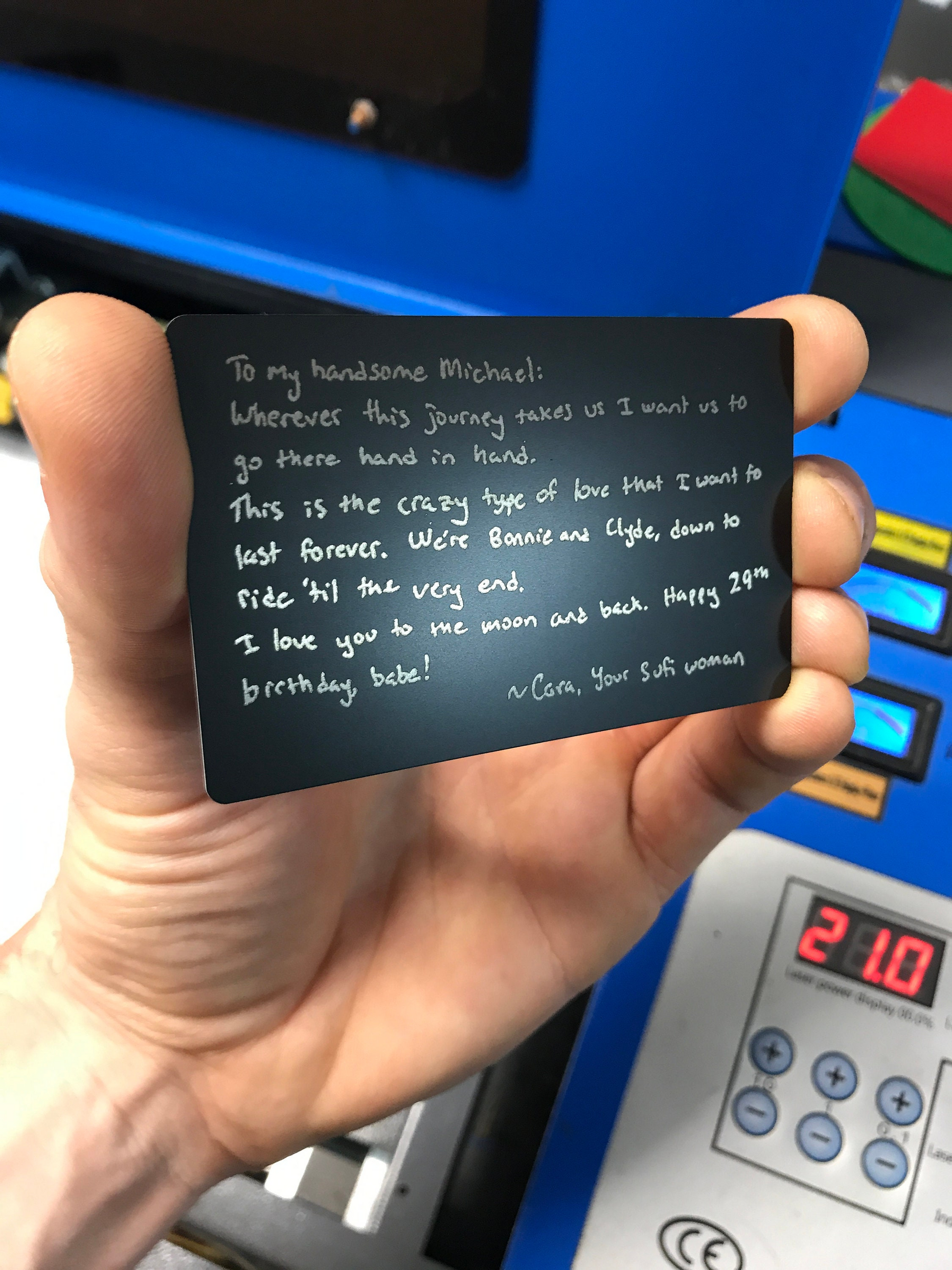 Hand holding a small black card with a heartfelt handwritten romantic message in front of a blue machine.