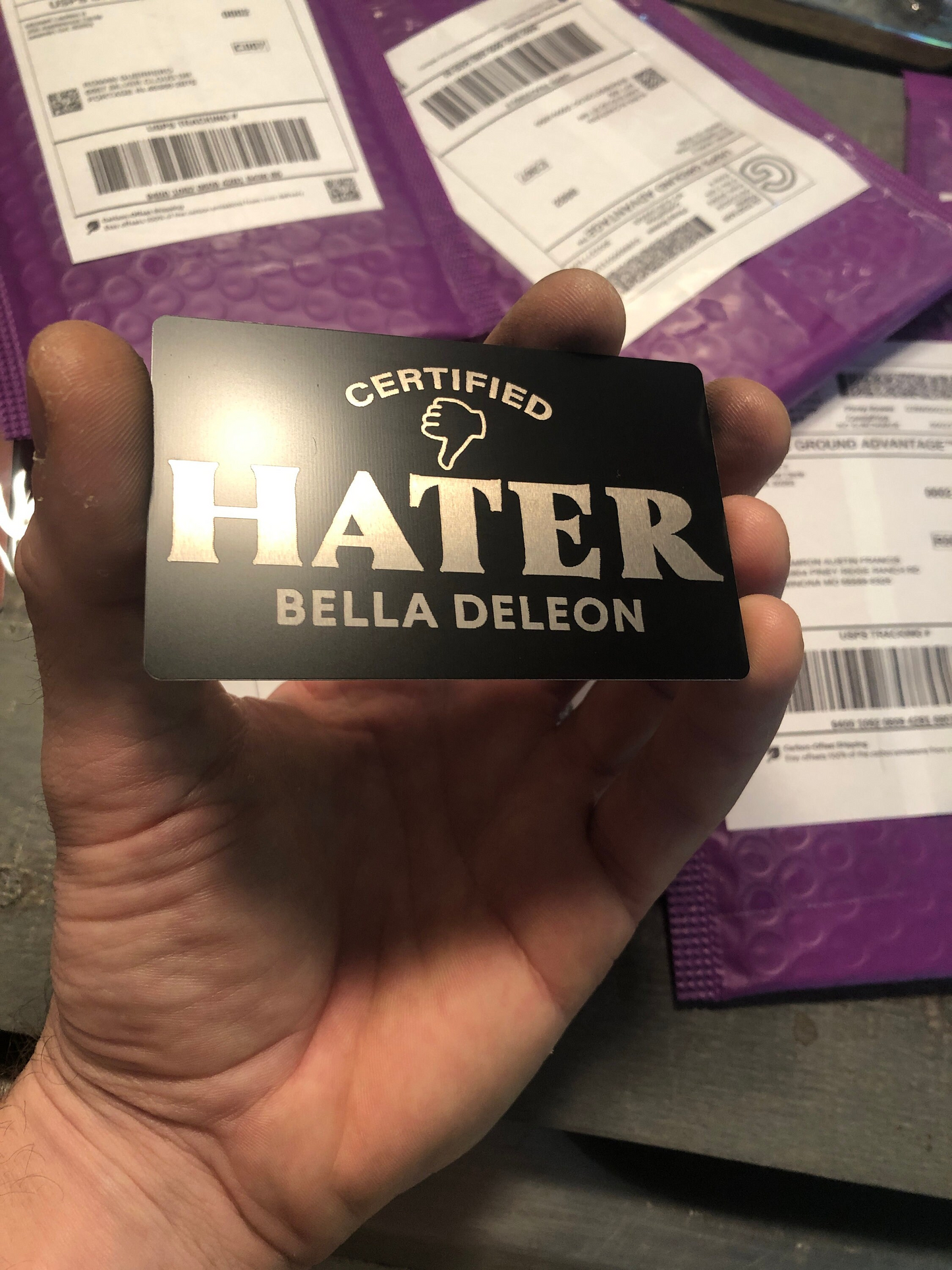 Hand holding a card with 'Certified Hater' text with custom name -- and a thumbs-down icon, with a purple background of mailers.