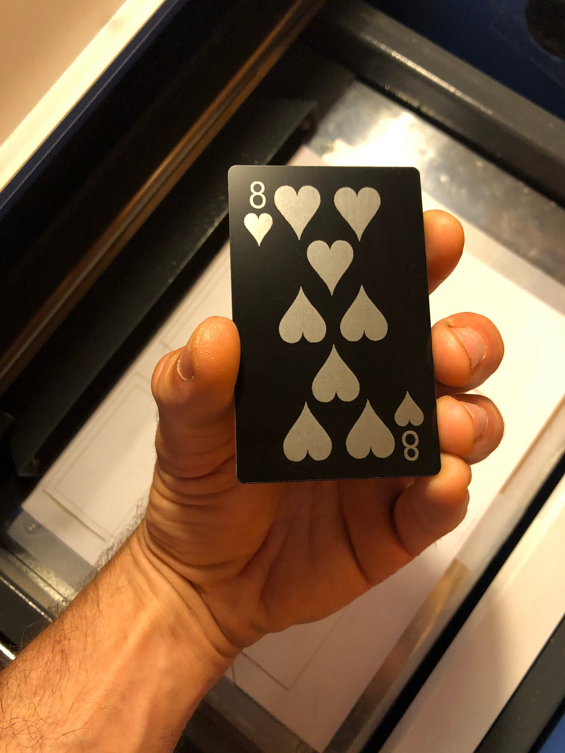 Hand holding an eight of hearts laser engraved playing card in front of a blurred machine background