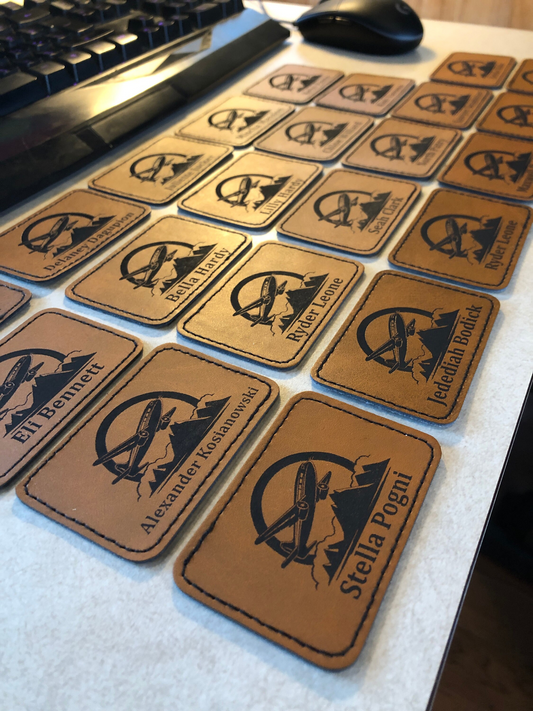 Set of brown leather coasters for a flight school with engraved student names and logo designs on a desk with a keyboard and mouse.