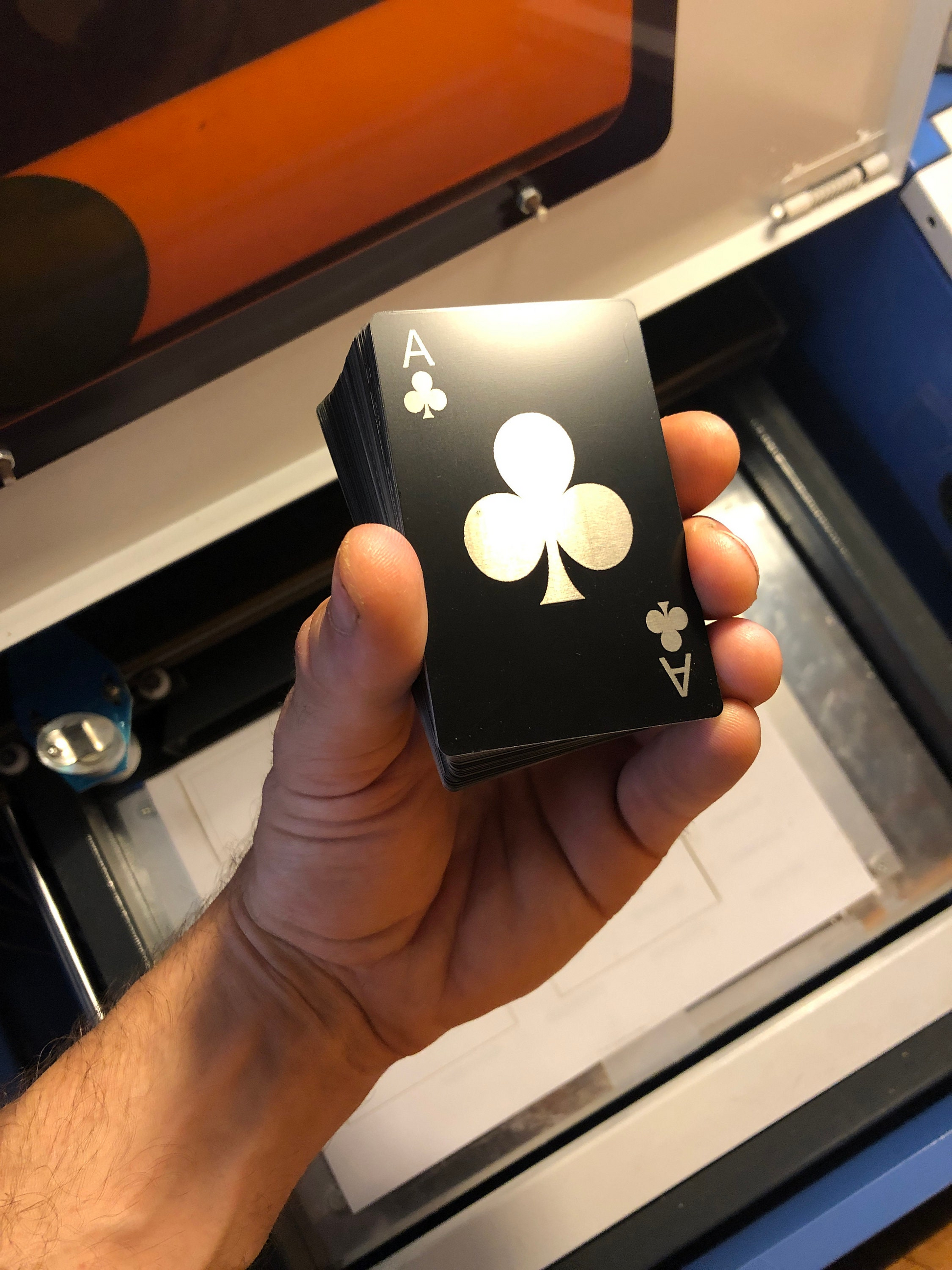Hand holding a deck of black metal playing cards with a ace of clubs on top in front of a laser engraving machine in the background.
