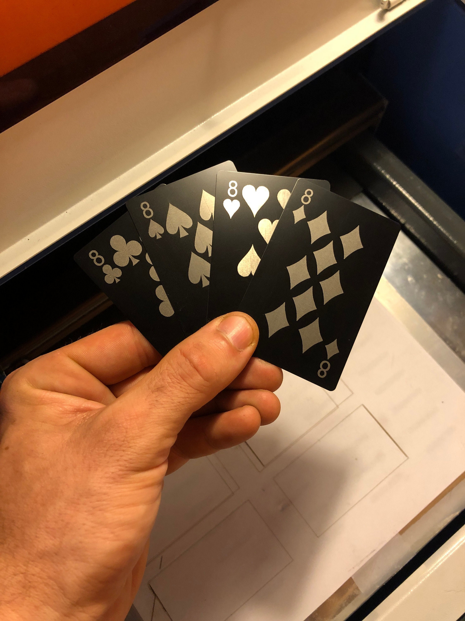hand holding four metal playing cards with Eight of Clubs, Eight of Spades, Eight of Hearts & Eight of Diamonds laser engraved in matte black/silver in front of a machine