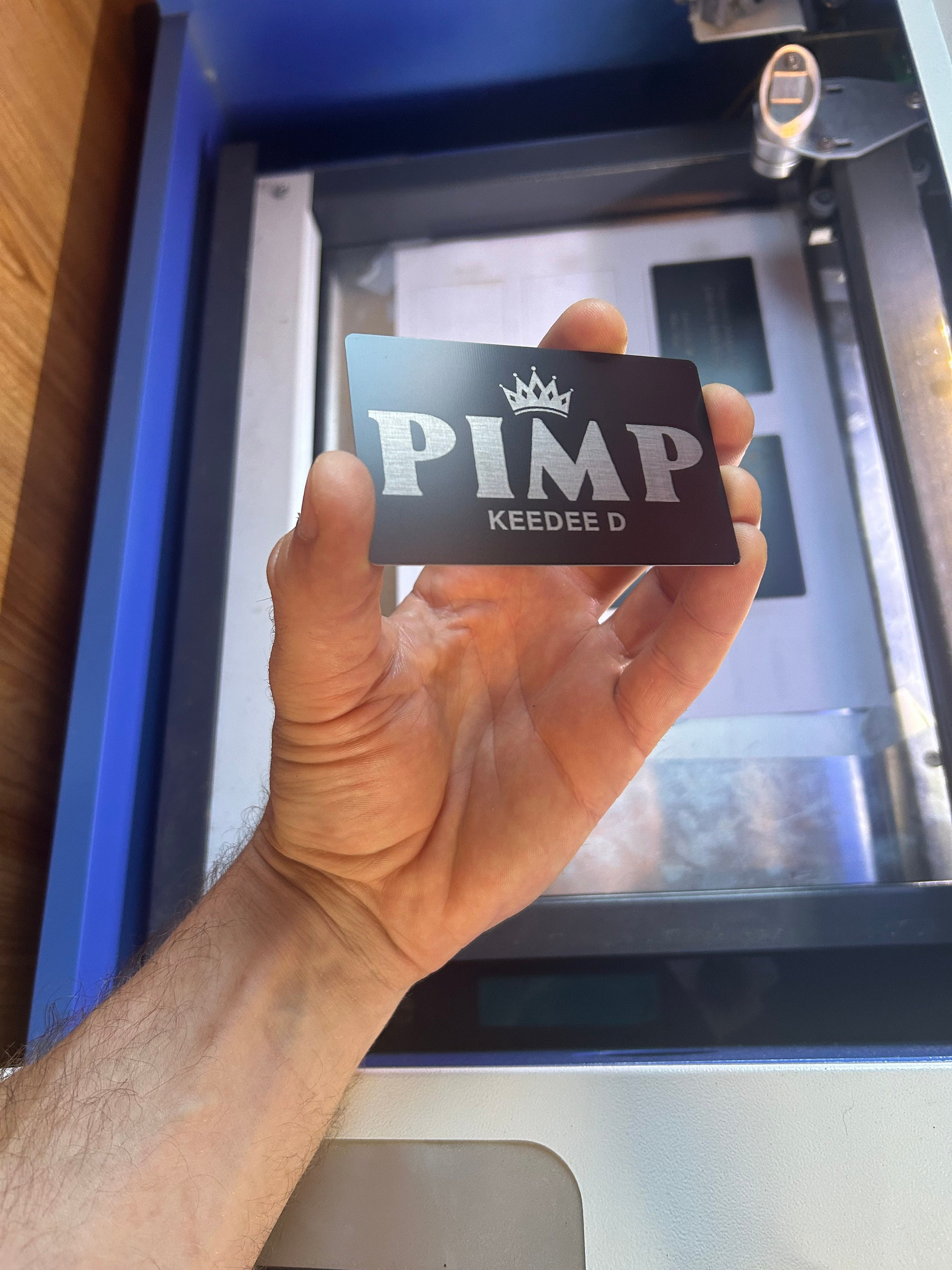 Hand holding a metal laser engraved novelty gift card with 'PIMP' and 'KEEDEE D' personalized text in front of a machine.
