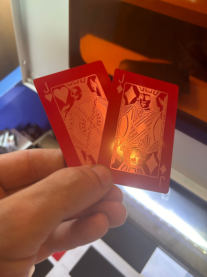 Two red/silver metal laser engraved playing cards, Jack of Hearts and Jack of Diamonds, held by a hand with a blurred background.