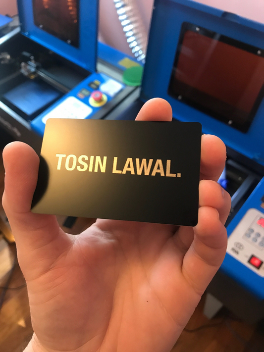 Hand holding a black card with 'first and last name' in silver laser engraved letters, with a blue machine in the background.