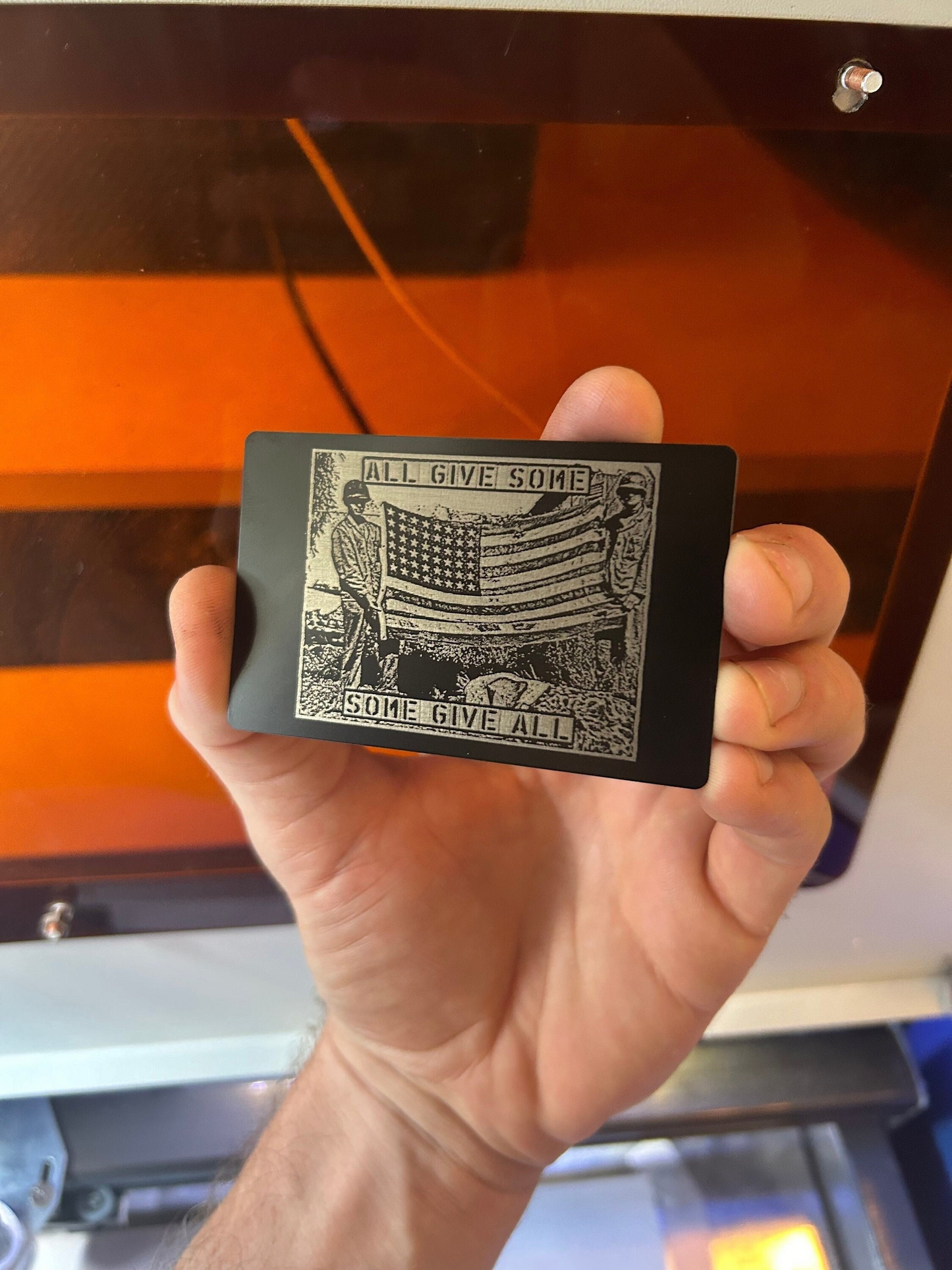 Hand holding a metal laser engraved veteran keepsake card with a graphic design of an American flag and All Give Some, Some Give All text.