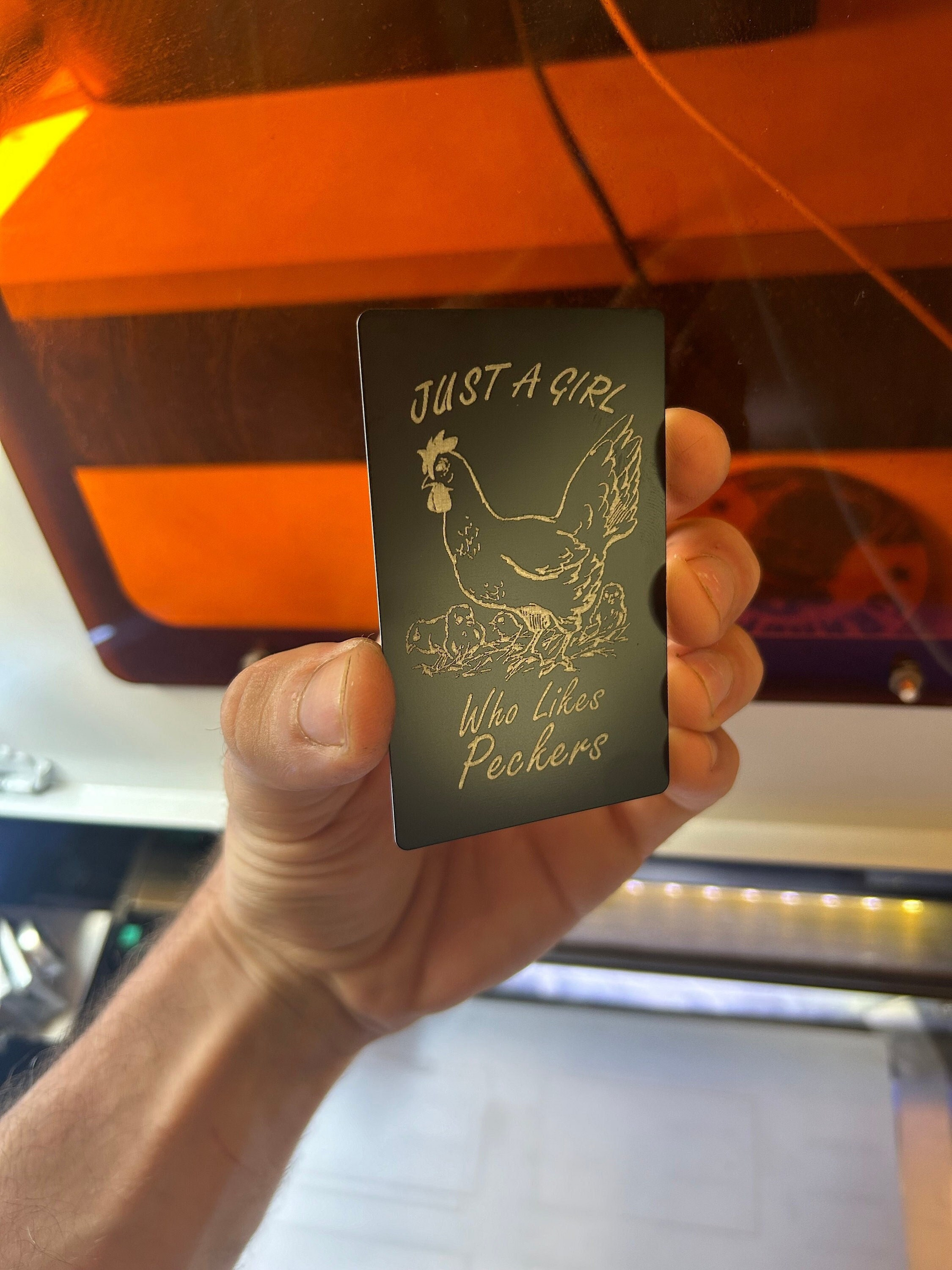 Hand holding a laser engraved black/silver metal friendsigving gag gifts for her card with Just A Girl Who Likes Peckers text and a chicken illustration 