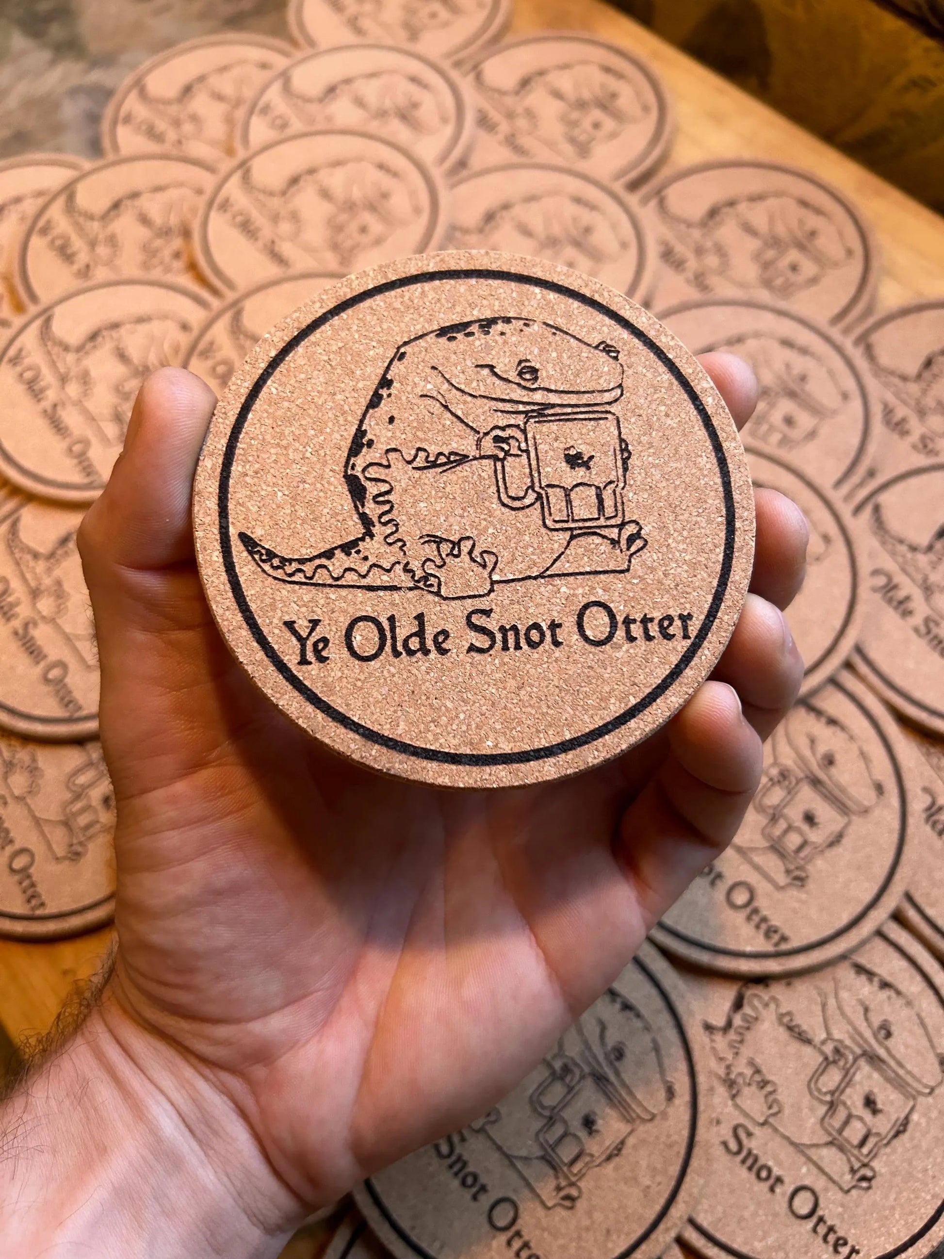 Coaster with 'Ye Olde Snot Otter' design held by a hand, surrounded by more coasters on a wooden surface.
