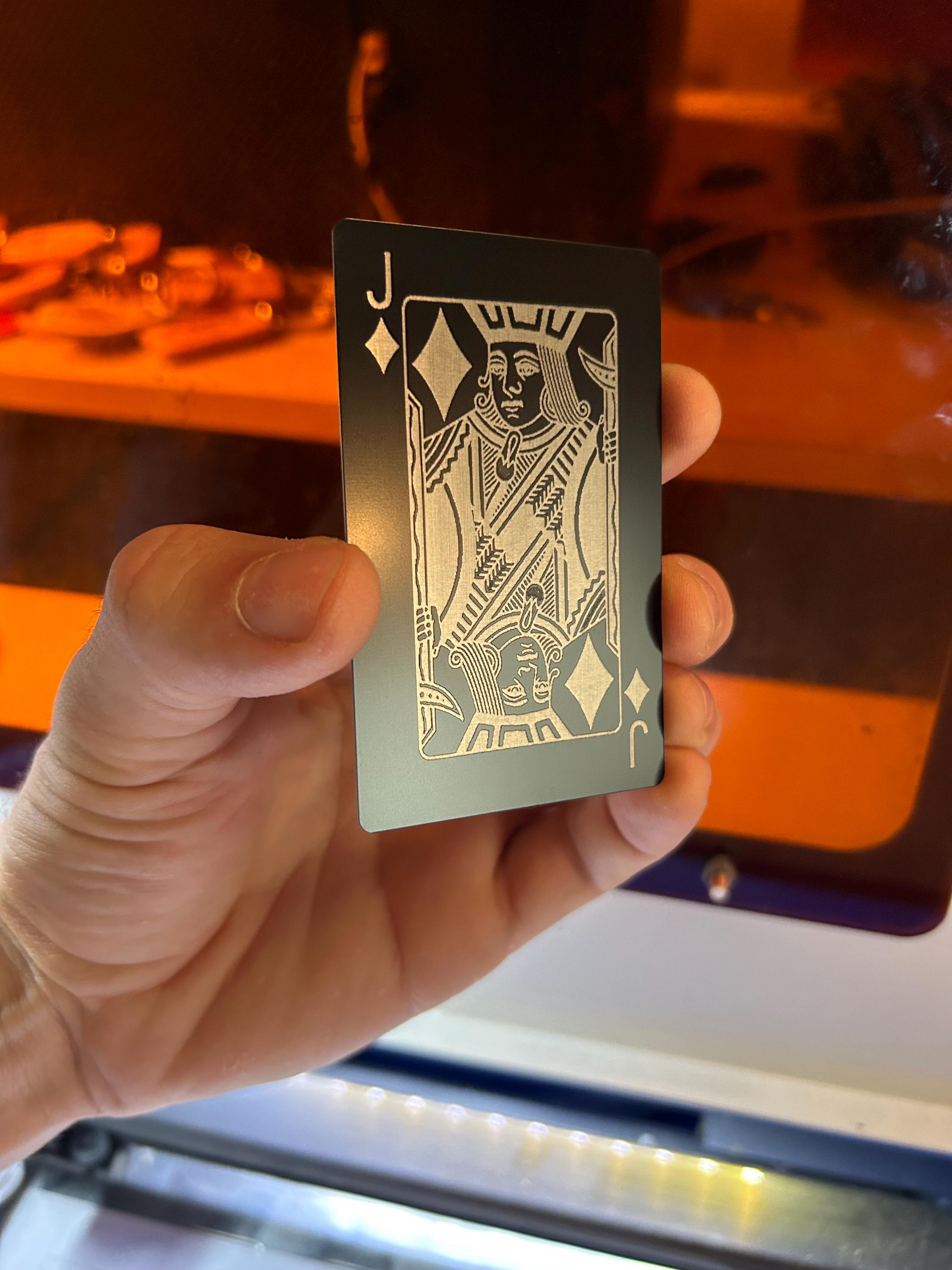 Hand holding a metal Jack of Diamonds playing card laser engraved in Black/Silver anodized aluminum, in front of machine