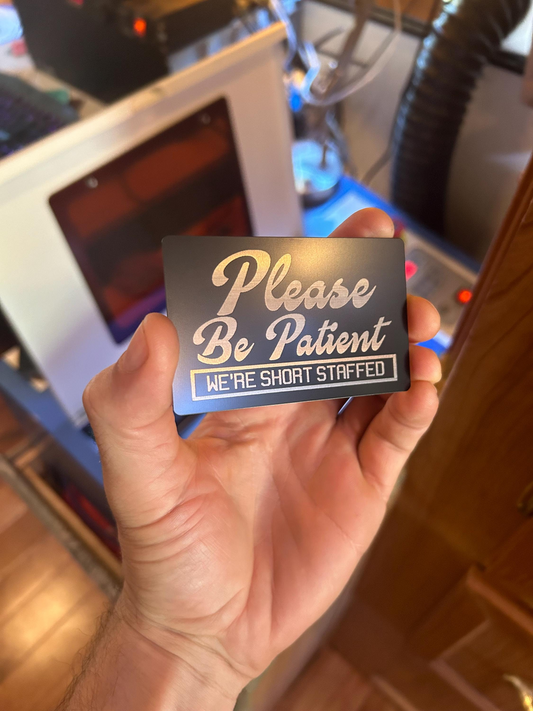 Hand holding a mini sign with 'Please Be Patient, We're Short Staffed' text in front of a blurred indoor background.