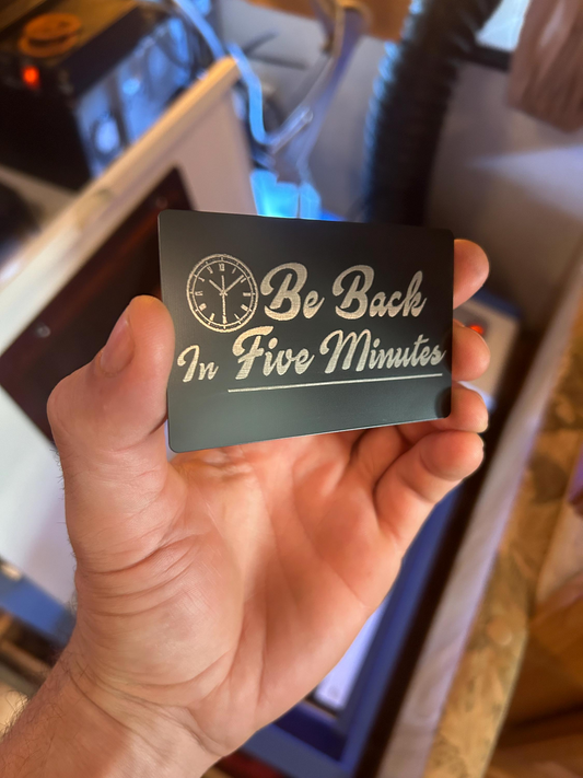 Hand holding a metal black card with 'Be Back In Five Minutes' text and clock design.