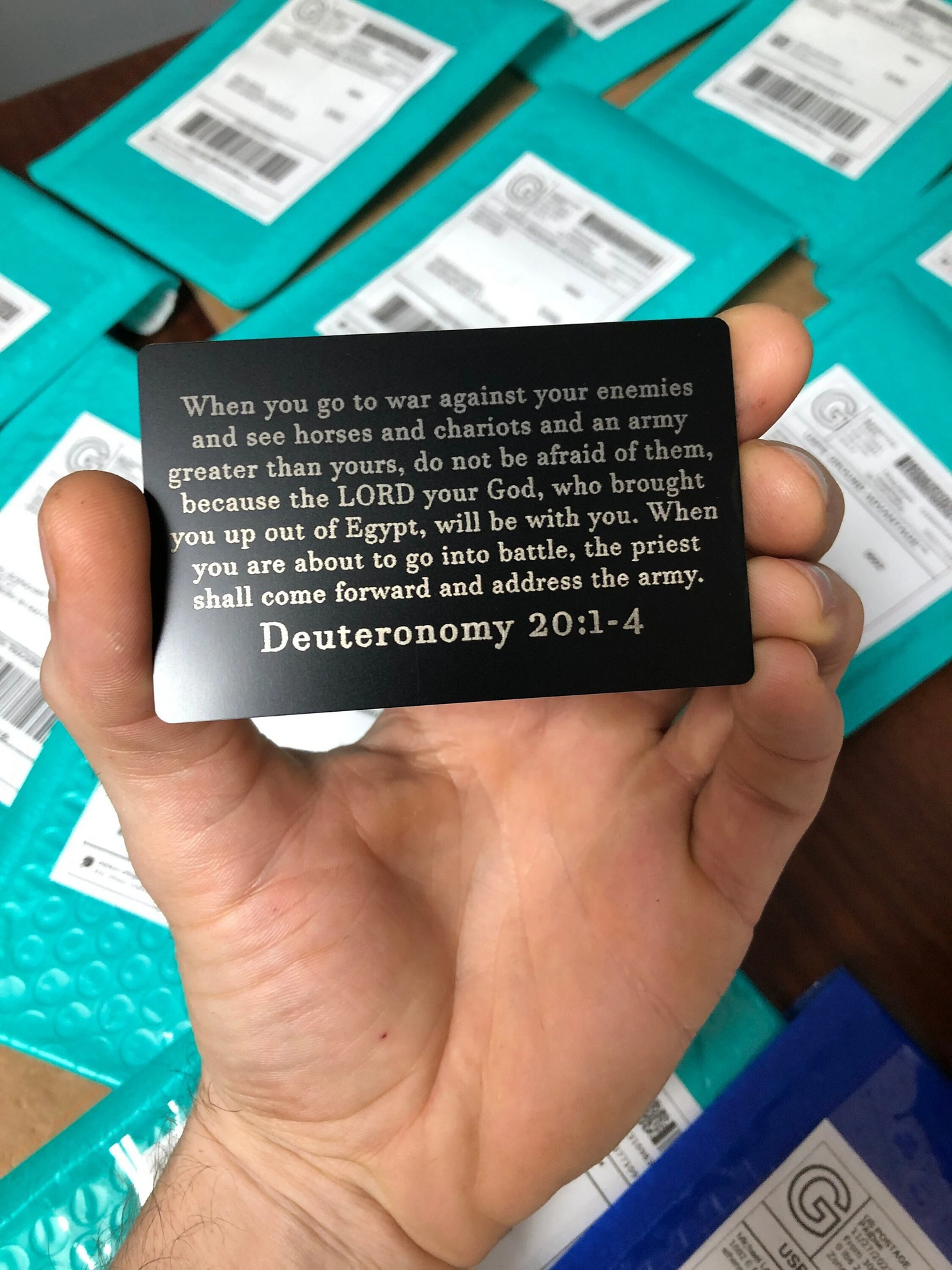 Hand holding a metal bible verse card with Deuteronomy 20:1-4 text in front of teal packaging