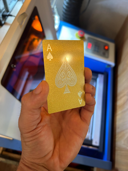 Hand holding a gold anodized aluminum metal playing card with a customized ace of spades design in front of a blurred background.