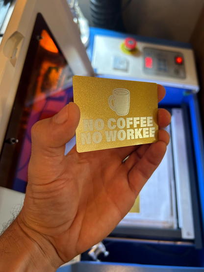 Hand holding a gold and silver metal business card with 'No Coffee No Workee' customized text and coffee cup graphic in front of a cnc machine.