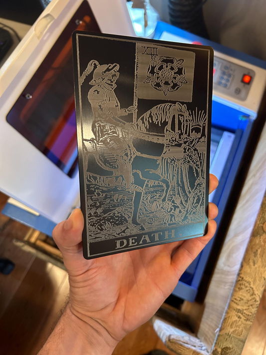 Hand holding a 5x7 oversized metal tarot card with "Death" design in matte black/silver, in front of a laser engraving machine