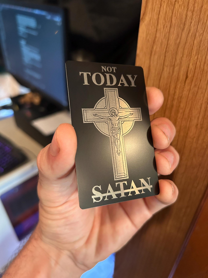 Hand holding a metal engraved card with a cross design and text 'Not Today Satan' in front of a blurred background.