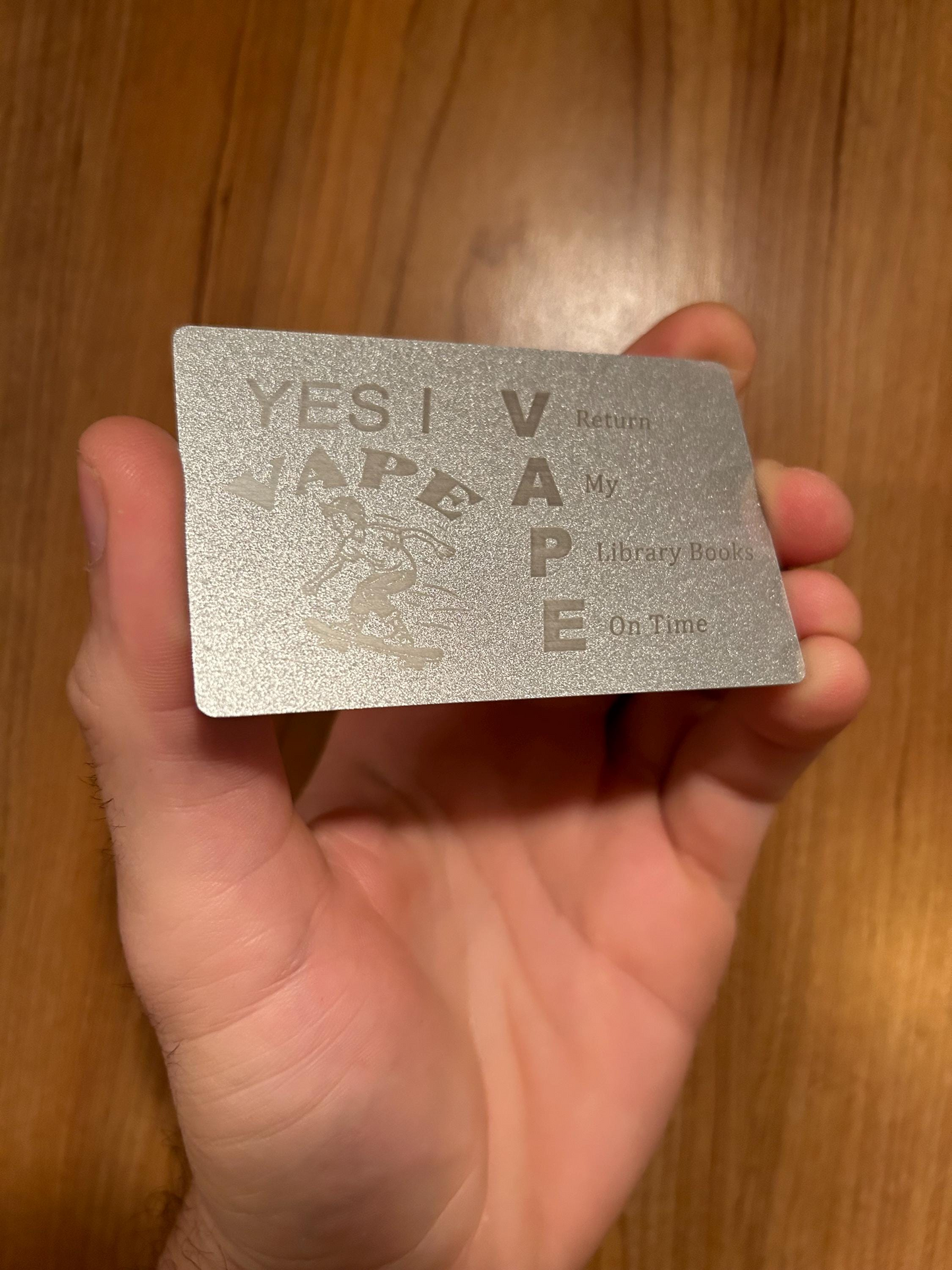 Hand holding a silver and silver laser engraved metal business card with text and graphics in front of a wooden surface