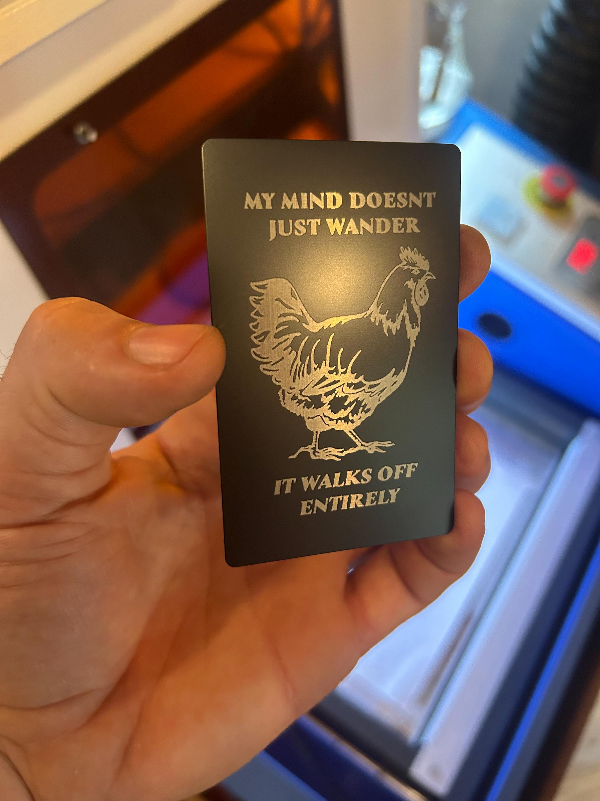 Hand showcasing a laser cut novelty metal card with engraved “My Mind Doesn’t Just Wander, It Walks Off Entirely” and chicken artwork