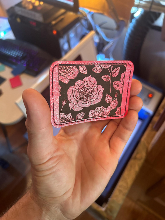 Hand holding a red patch with laser engraved rose and floral design in front of a laser engraving machine background