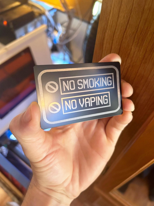 Hand holding a 'No Smoking No Vaping' sign in front of a blurred indoor background.