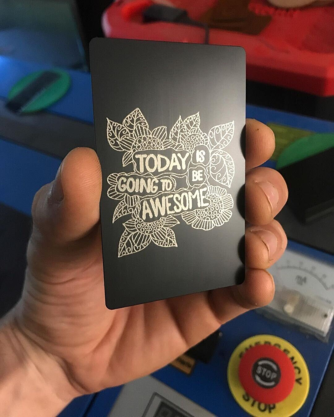 Hand holding a black metal keepsake card with silver laser engraved "Today Is Going To Be Awesome" text and floral pattern design, set against a colorful background.