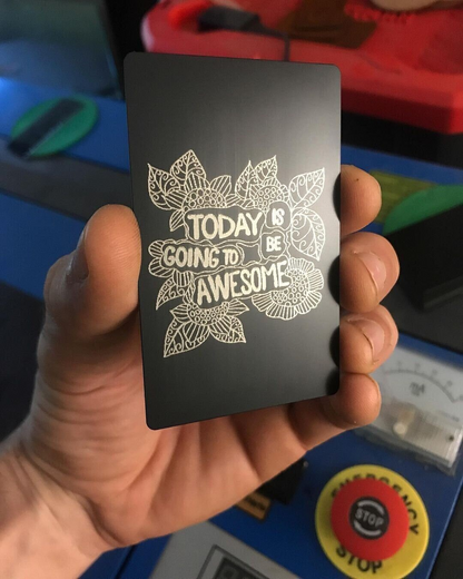 Hand holding a black metal keepsake card with silver laser engraved "Today Is Going To Be Awesome" text and floral pattern design, set against a colorful background.