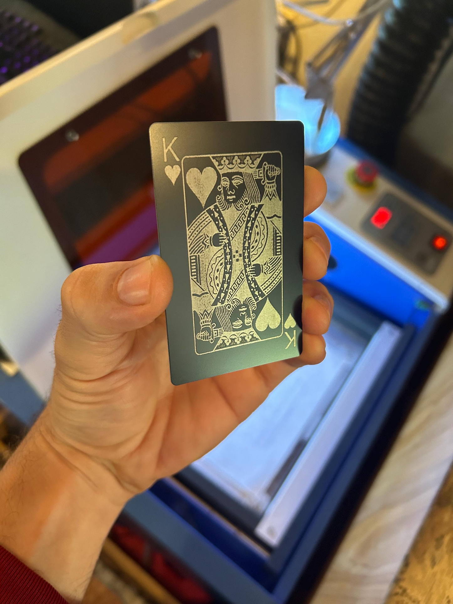 Hand holding a black playing card with a silver laser engraved king of hearts design in front of a machine.