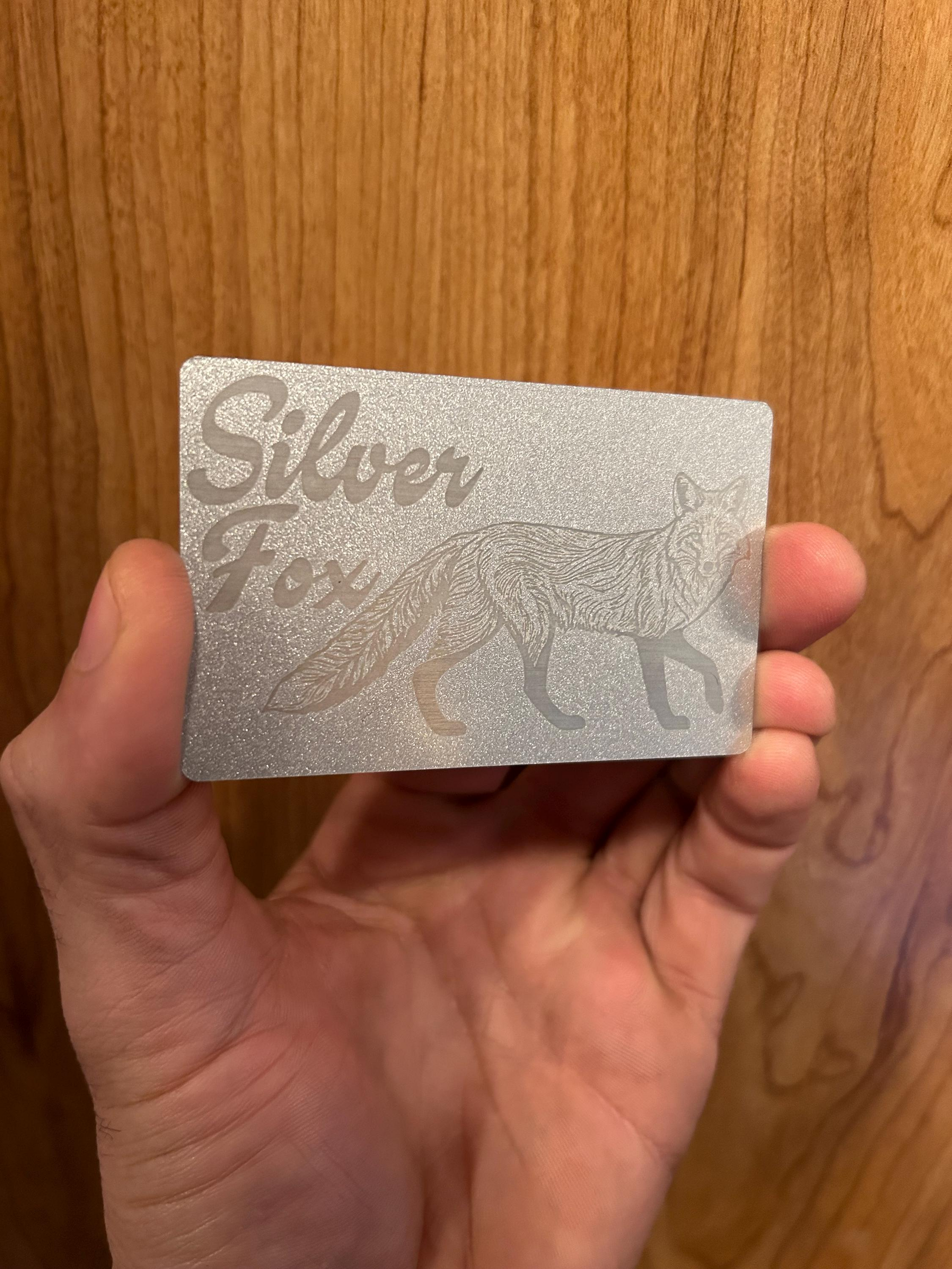 Hand holding a 'Silver Fox' metal novelty card against a wooden background