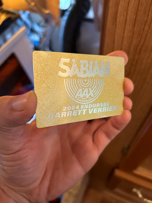 Hand holding a laser engraved metal business card with gold/silver logo and personalized text for endorsee at promotional event, shown in blurred indoor setting