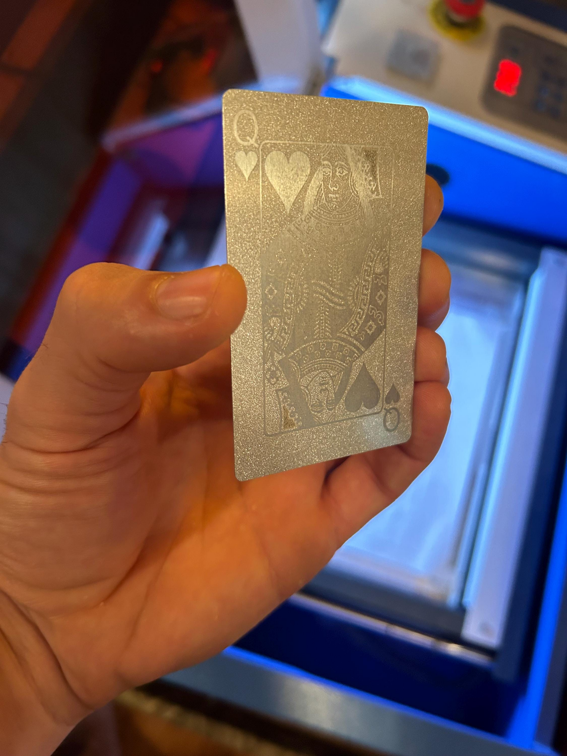Hand holding a silver/silver laser engraved metallic playing card with intricate Queen of Hearts design against a blurred background