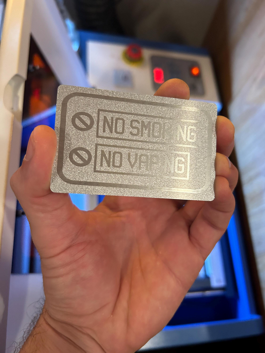 Hand holding a customized laser engraved metal business card signage placard 'No Smoking No Vaping' text in front of a blurred machine background.