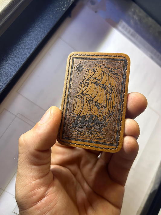 Hand holding a leather burned patch with a ship illustration on a light background