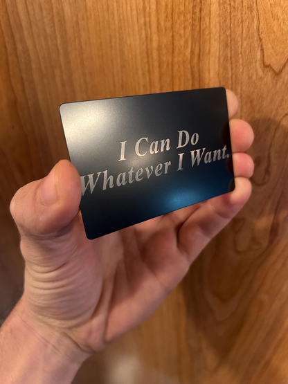 Hand holding a black metal keepsake card with 'I Can Do Whatever I Want' text against a wooden background