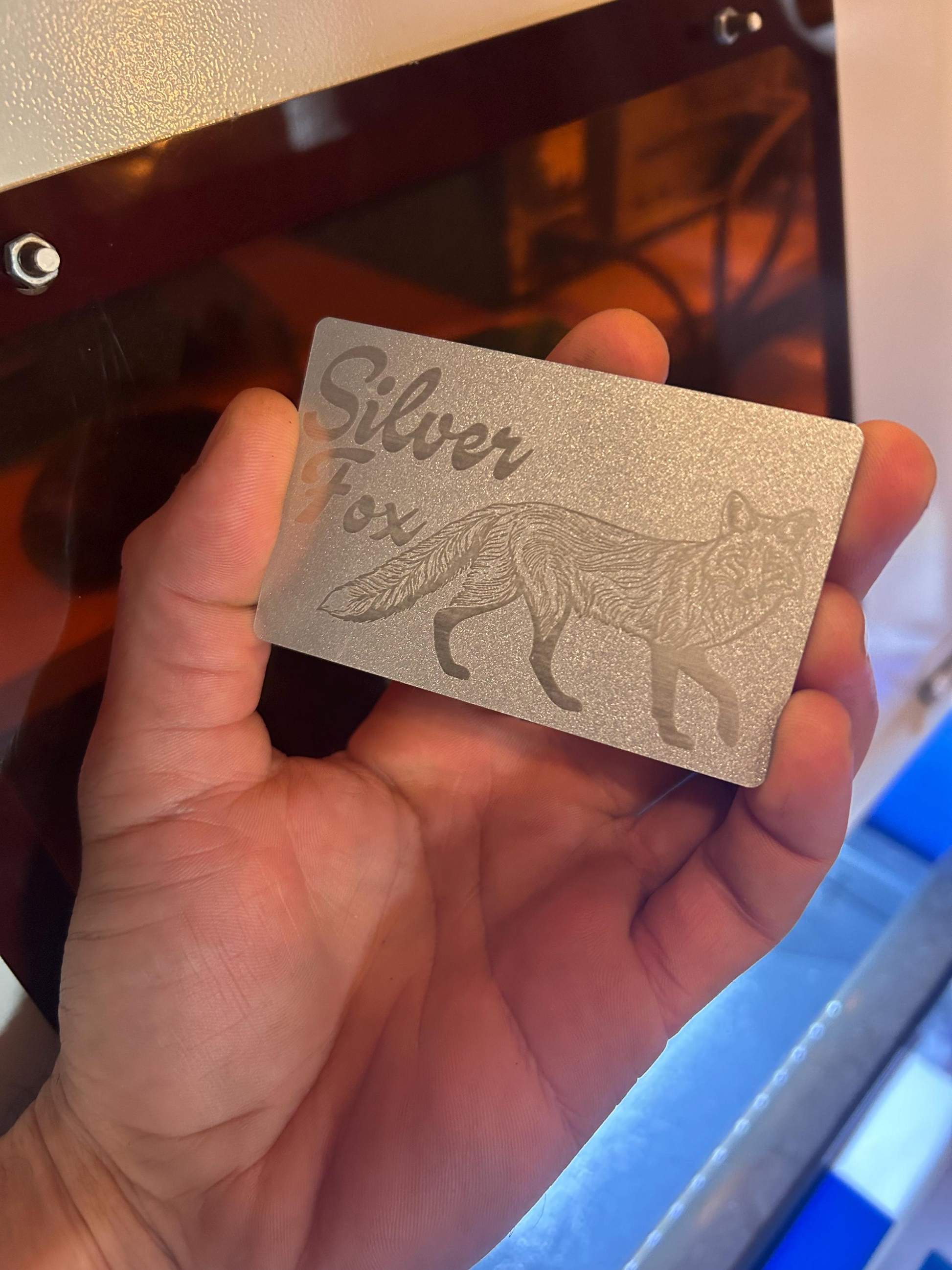 Hand holding a metal friendsgiving card with 'Silver Fox' engraving and fox graphic in front of a laser engraving machine .