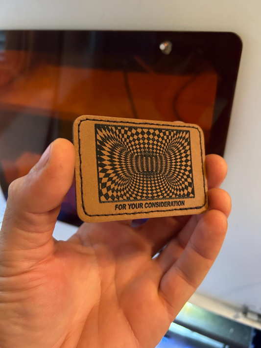 Hand holding a 'For Your Consideration' brown leather patch with a black and white optical illusion pattern.