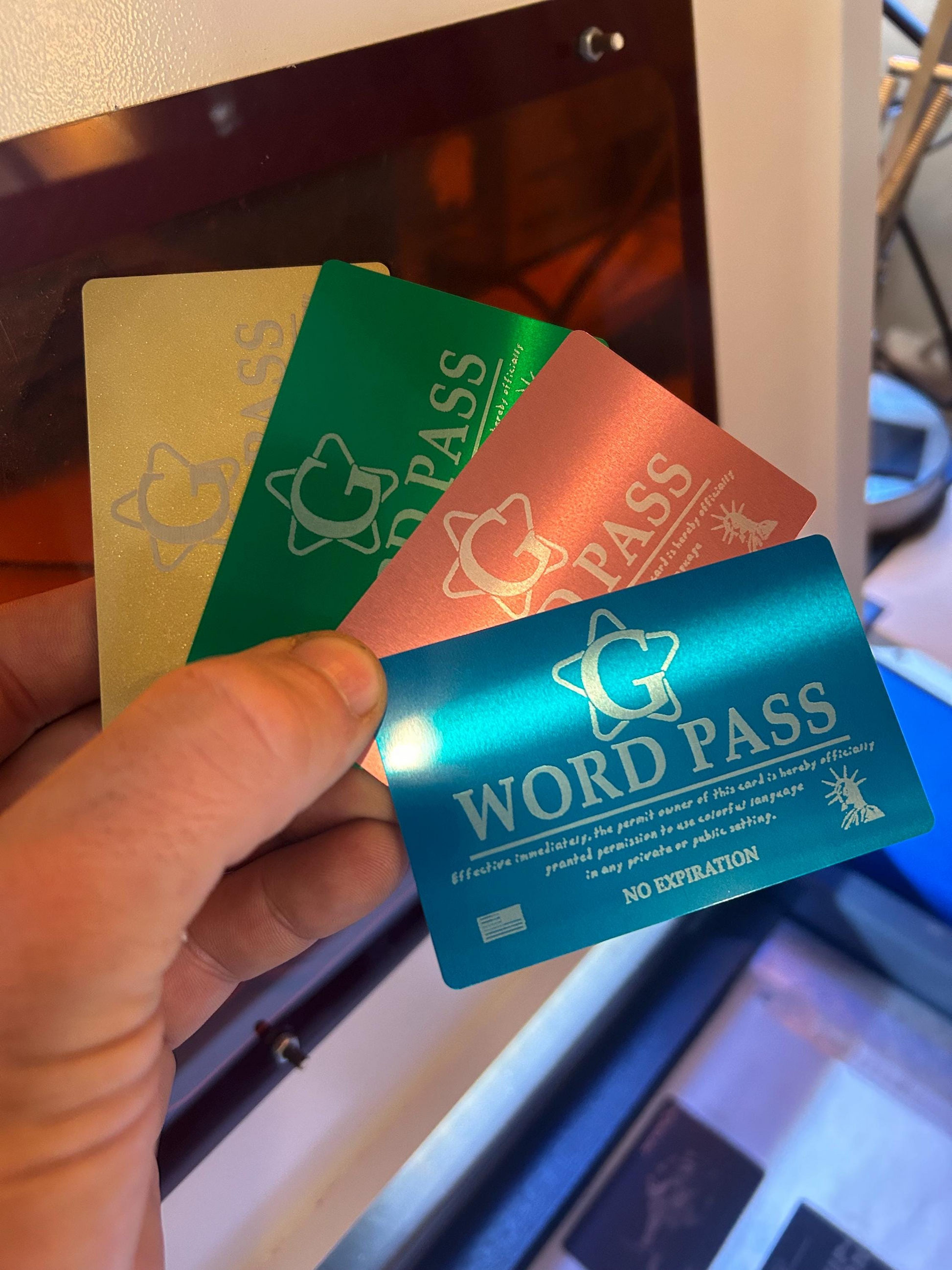 Hand holding a blue, rose gold, green, gold metal novelty gift cards with  'G WORD PASS' laser engraved on each  with a production laser engraving machine in the background