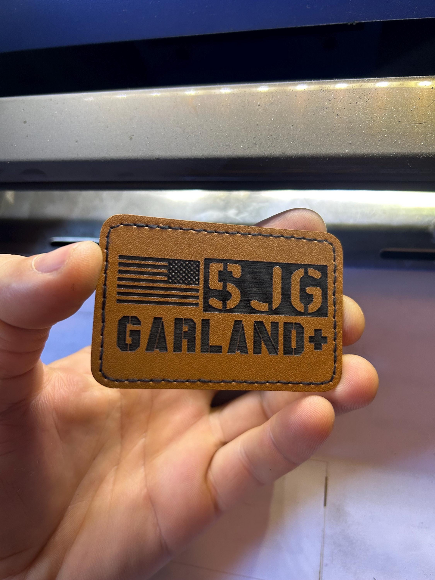 Brown leather patch with 'call sign and last name' text laser engraved, held in a hand against a machine background