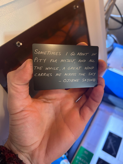 Hand holding a black card with handwritten ojibwe quote text engraved in an indoor setting