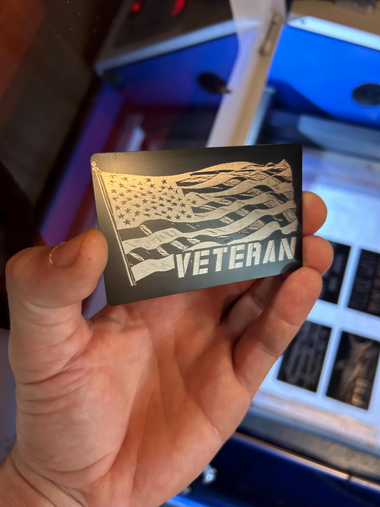 Hand holding a black metal wallet card with a silver engraved flag design and 'VETERAN' text against a blurred industrial background.