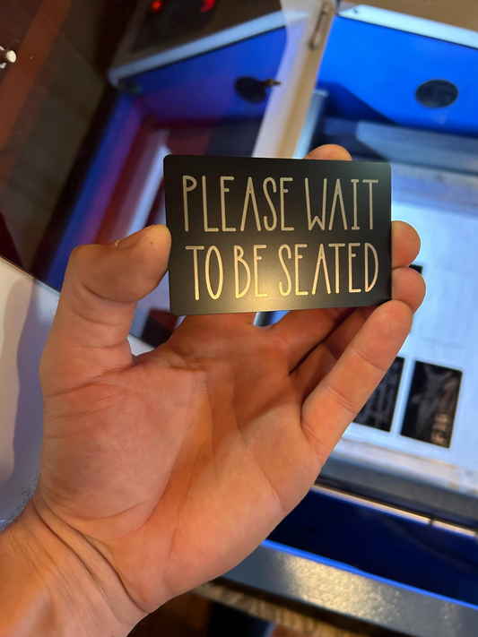 Hand holding a small metal placard with silver text 'PLEASE WAIT TO BE SEATED' in front of a blue machine.