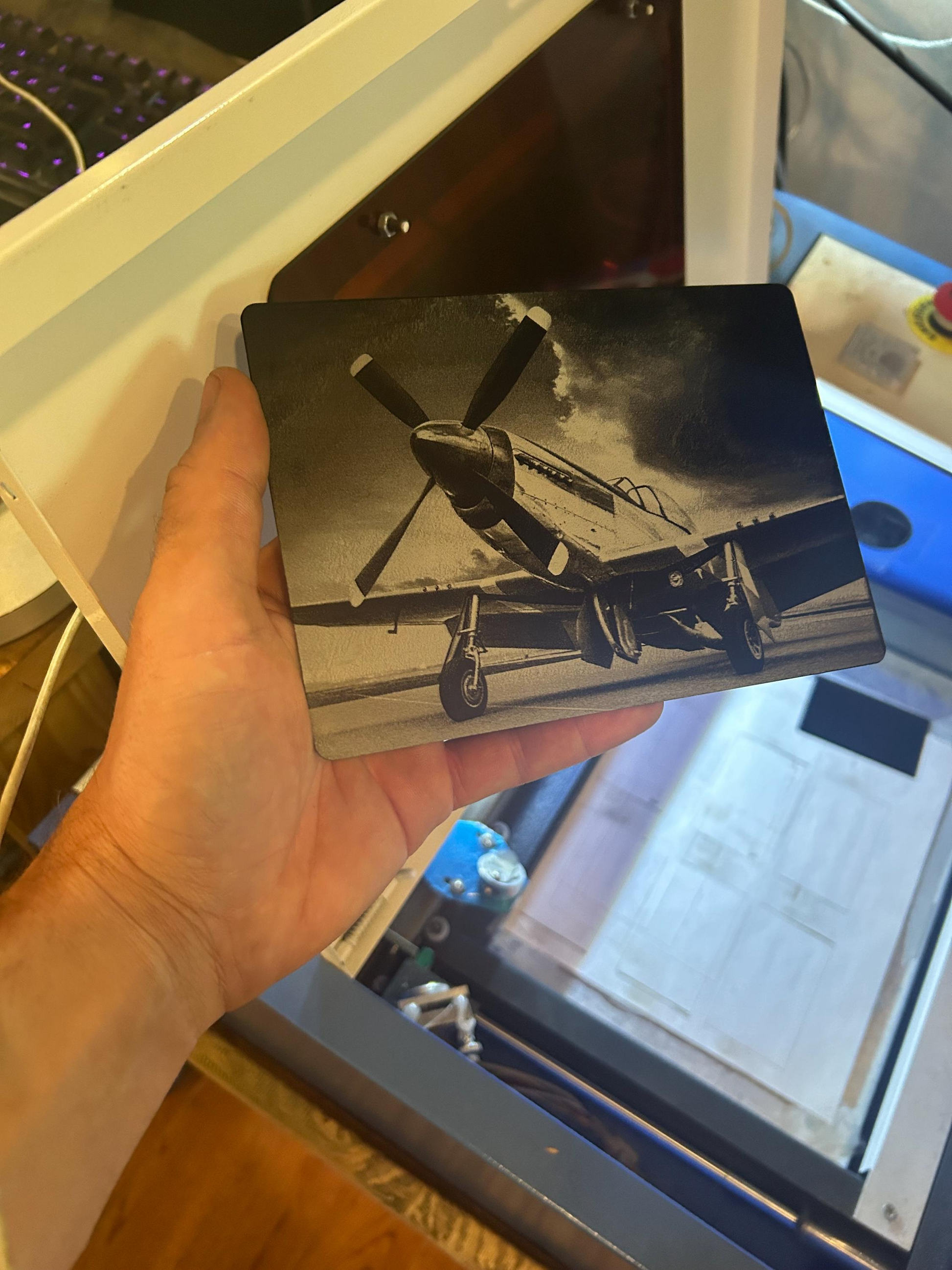 Hand holding a black and silver laser engraved P-51 plaque of an airplane in front of a computer screen.