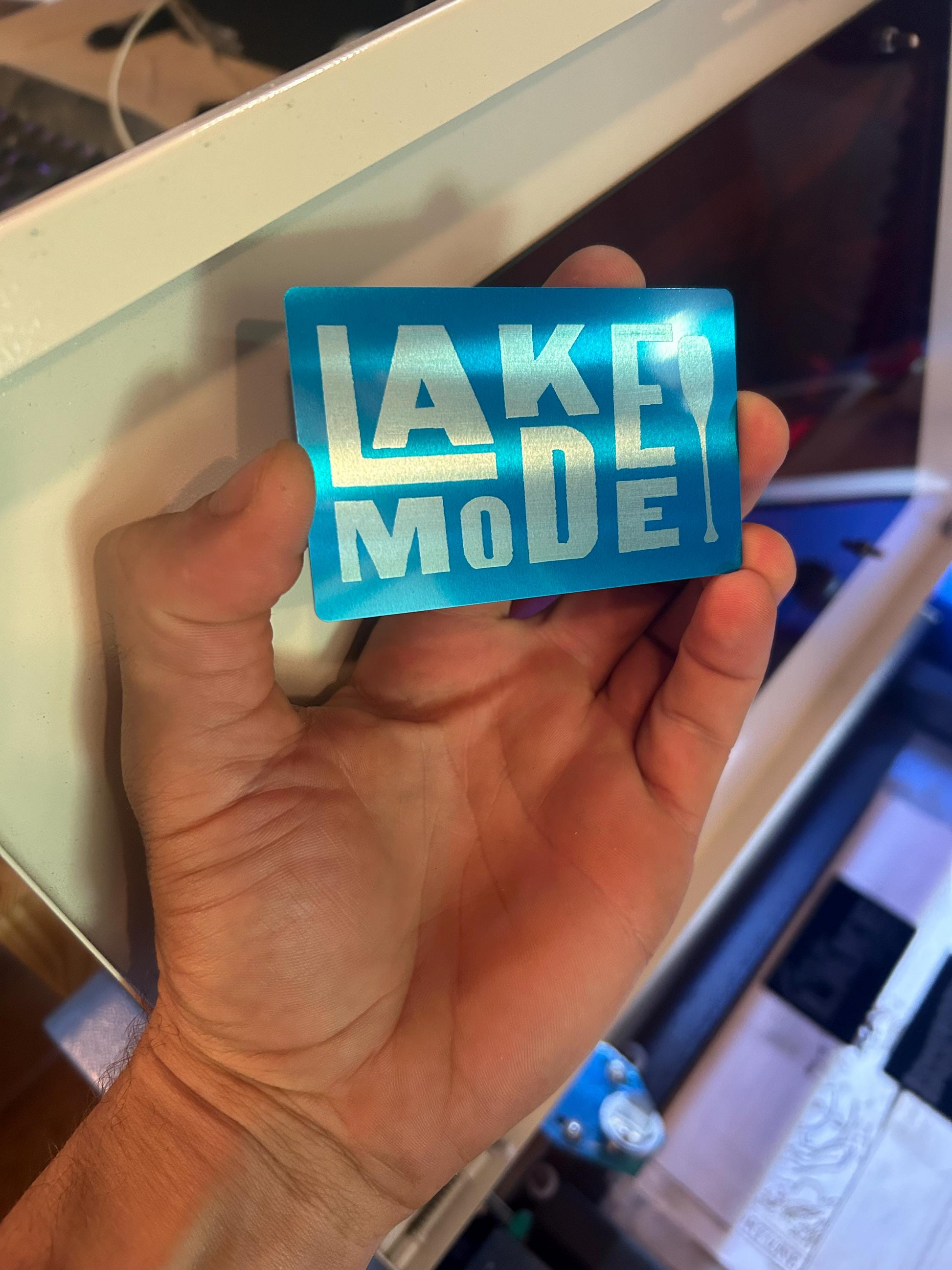 Angled shot of custom engraved metal lake gift card – “Lake Mode” typography with boat oar design in blue/silver