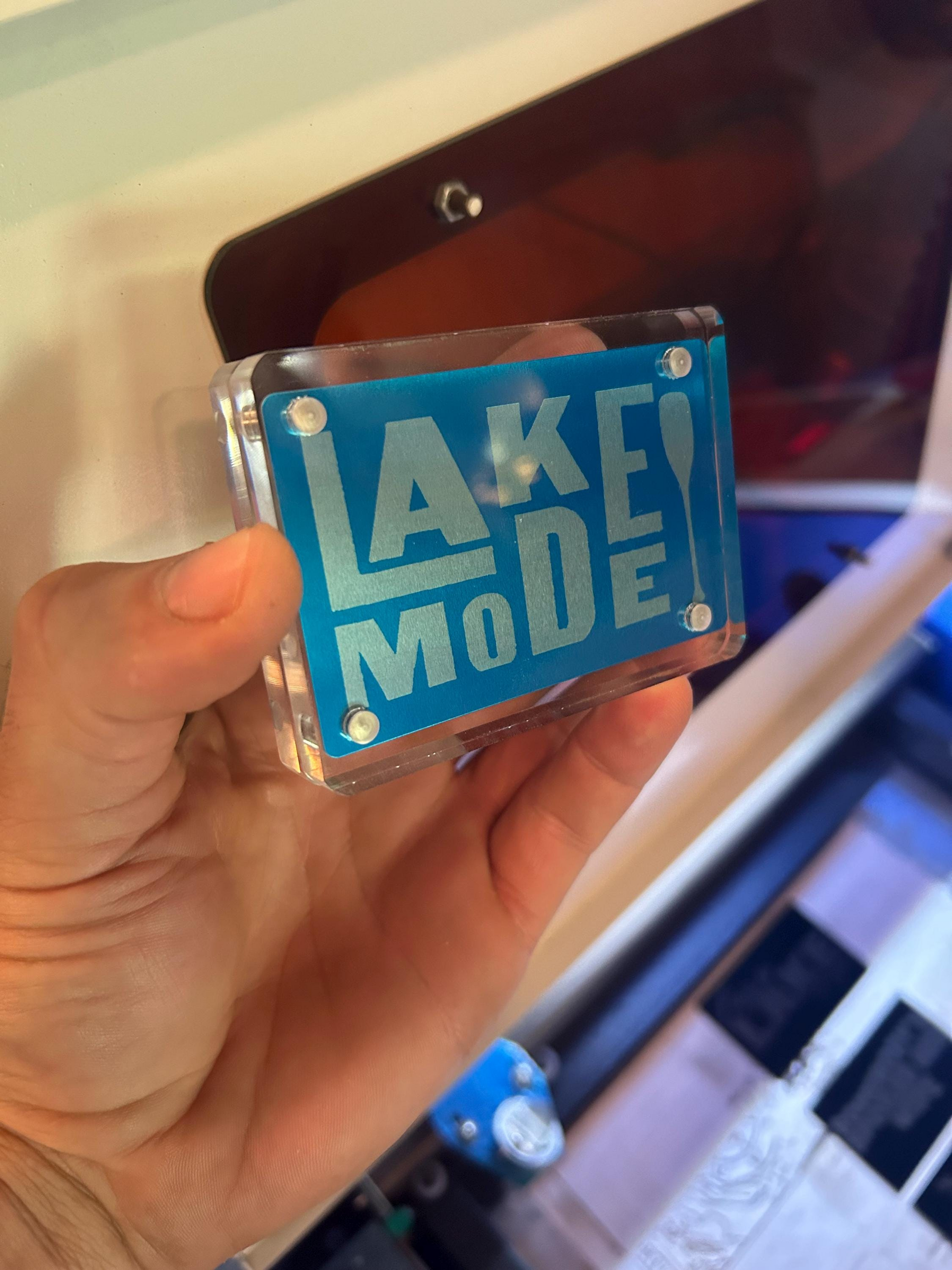 Hand holding a blue and silver metal keepsake card engraved with “Lake Mode” text and boat oar graphic
