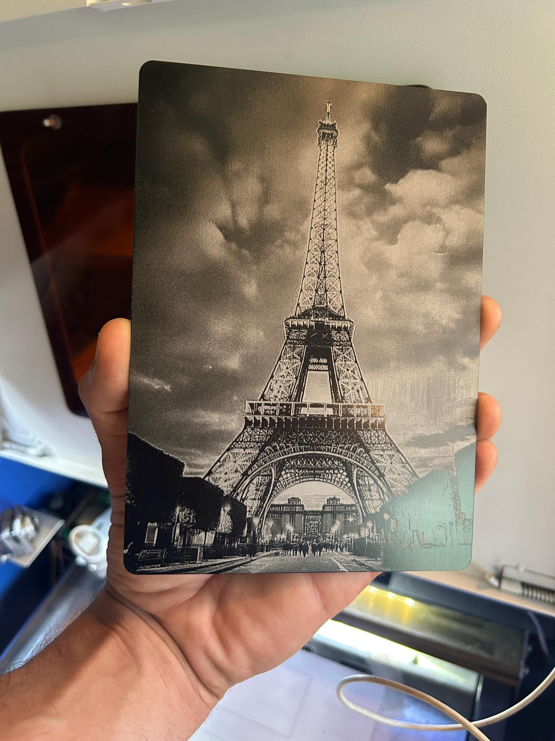 Hand holding a decorative plaque size 5x7" with a black and white highly detailed engraving of the Eiffel Tower against a cloudy sky.