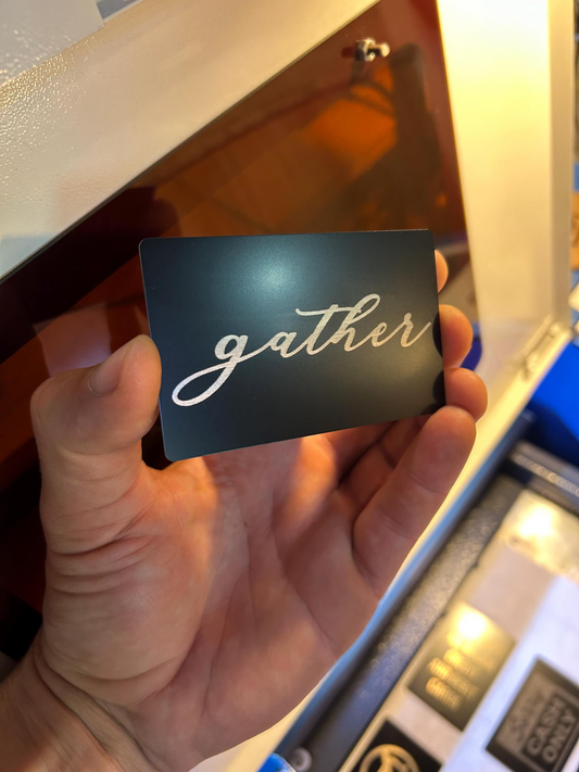 Angled view of custom engraved metal placard featuring the word “gather” in silver on black